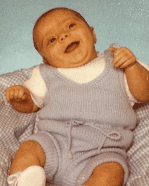 Brendan as a smiling baby in a pale-blue knit outfit.