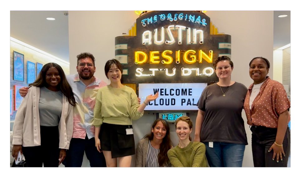 The IBM Cloud PAL design team gathered under a vintage 'Welcome Cloud PAL' marquee at the Austin Design Studio — seven smiling people of varied gender and ethnicity, posing under a backlit yellow-bulb neon sign.